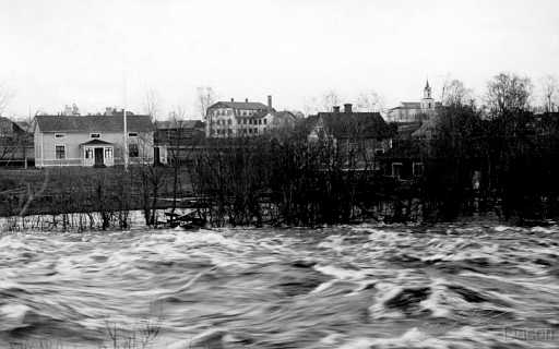 19xx Råneå älven kyrkan .jpg - Lundbergs hus på holmen har besök av vårfloden och den mäktiga Lundins skofabrik tronar i mitten med kyrkan i bakgrunden. 